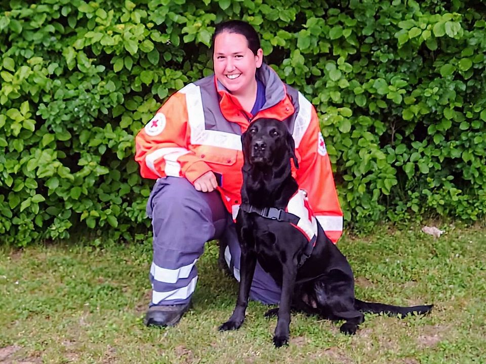 Lächelnde Rettungshundeführerin Saskia in orange-grauer Einsatzkleidung kniet auf einer Wiese neben einem sitzenden, schwarzen Rettungshund Cayuki mit Geschirr; im Hintergrund dichter grüner Busch.