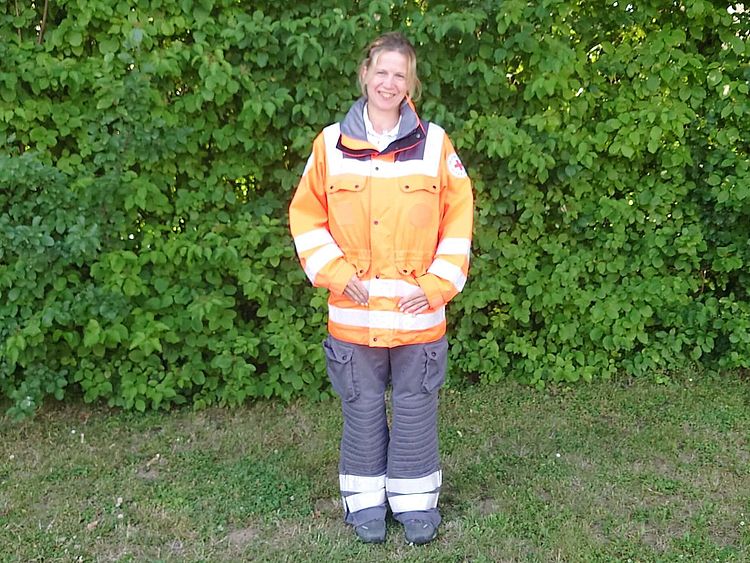 Lächelnde Helferin Janine in orange-grauer Einsatzkleidung steht auf einer Wiese; im Hintergrund dichter grüner Busch.