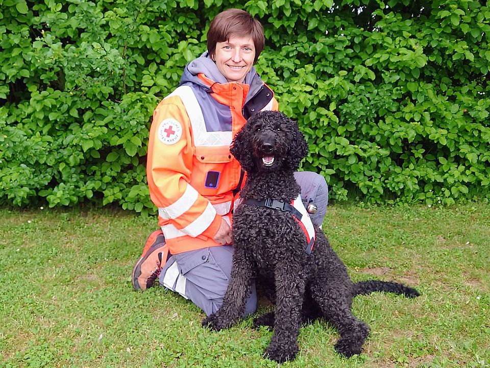 Lächelnde Rettungshundeführerin Elisabeth in orange-grauer Einsatzkleidung kniet auf einer Wiese neben einem sitzenden, schwarzen Rettungshund Kiko mit Geschirr; im Hintergrund dichter grüner Busch.