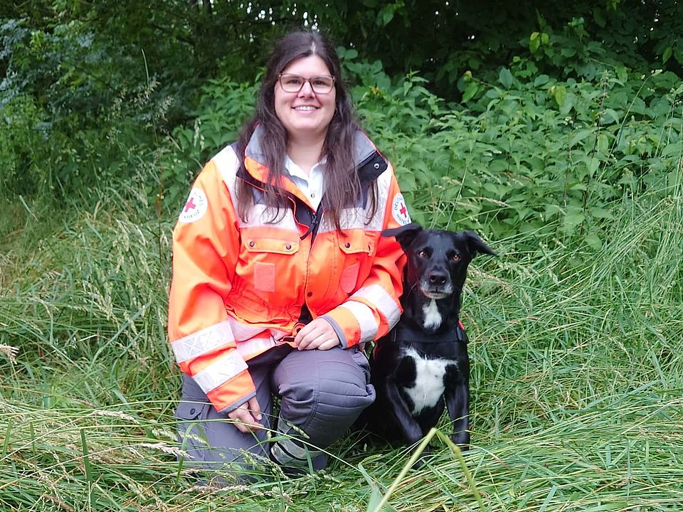 Lächelnde Rettungshundeführerin Ann-Kathrin in orange-grauer Einsatzkleidung kniet auf einer Wiese neben sitzendem, schwarz-weißen Rettungshund Pepe mit Geschirr; im Hintergrund dichter grüner Busch.