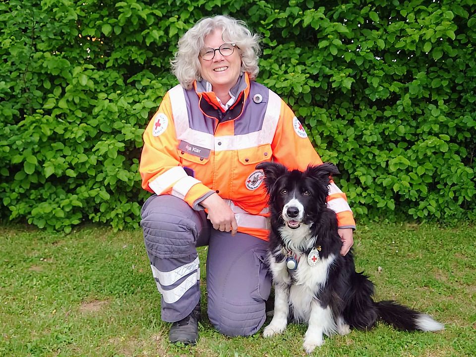 Lächelnde Rettungshundeführerin Anja in orange-grauer Einsatzkleidung kniet auf einer Wiese neben einem sitzenden, schwarz-weißen Rettungshund Mascha mit Geschirr; im Hintergrund dichter grüner Busch.