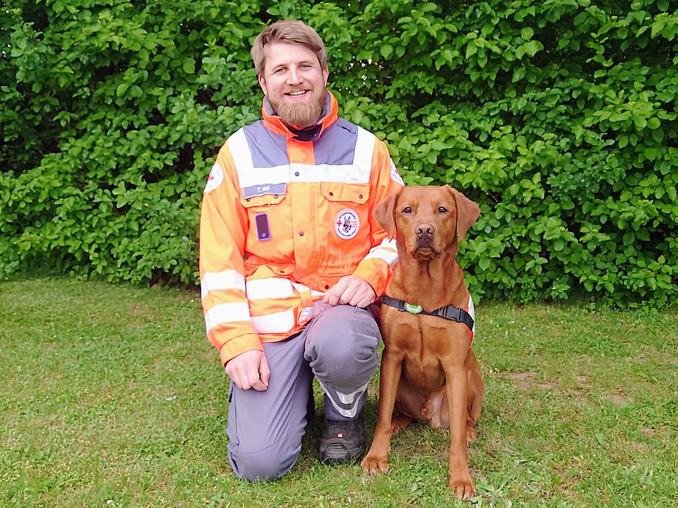 Lächelnder Rettungshundeführer Tobie in orange-grauer Einsatzkleidung kniet auf einer Wiese neben einem sitzenden, braunen Rettungshund Spike mit Geschirr; im Hintergrund dichter grüner Busch.