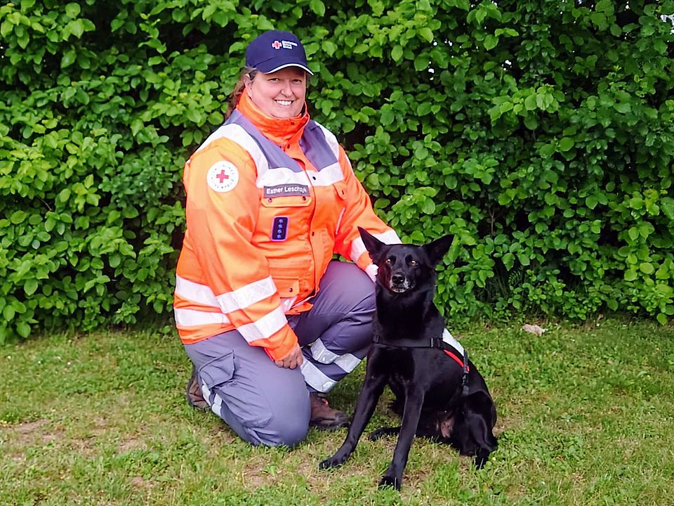 Lächelnde Rettungshundeführerin Esther in orange-grauer Einsatzkleidung kniet auf einer Wiese neben einem sitzenden, schwarzen Rettungshund Suki mit Geschirr; im Hintergrund dichter grüner Busch.