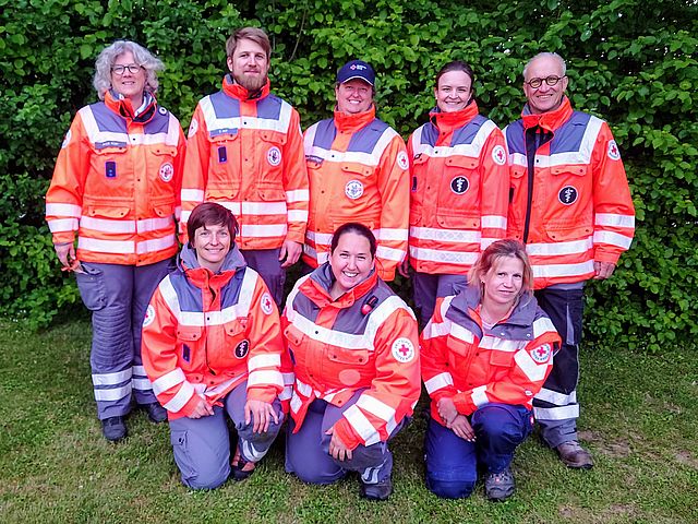 Gruppenfoto der Mitglieder der DRK Rettungshundestaffel Friedberg-Wöllstadt in Einsatzkleidung, lächelnd vor einer grünen Hecke.