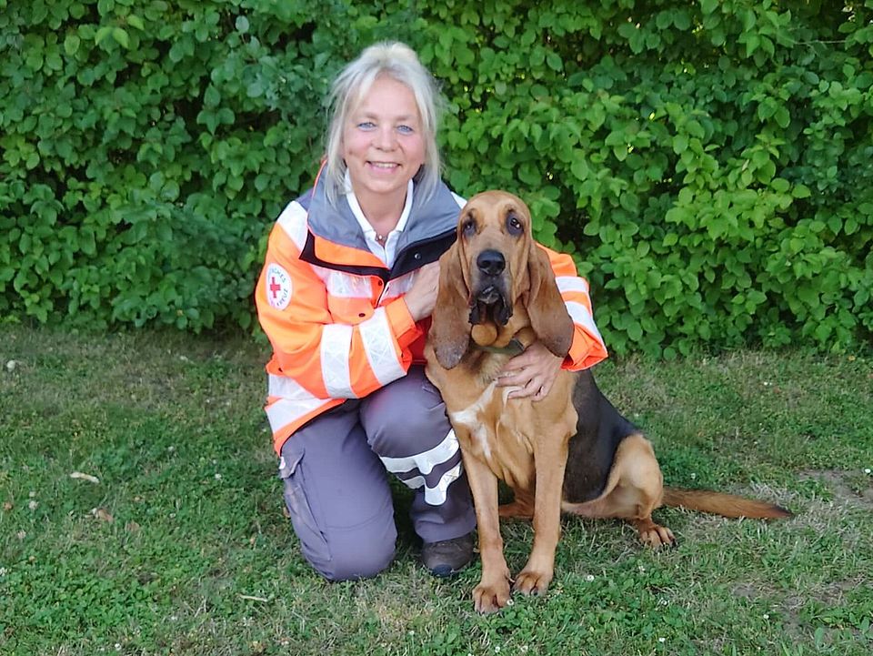 Lächelnde Rettungshundeführerin Christine in orange-grauer Einsatzkleidung kniet auf einer Wiese neben sitzendem, braun-schwarzem Rettungshund Piccolina; im Hintergrund dichter grüner Busch.