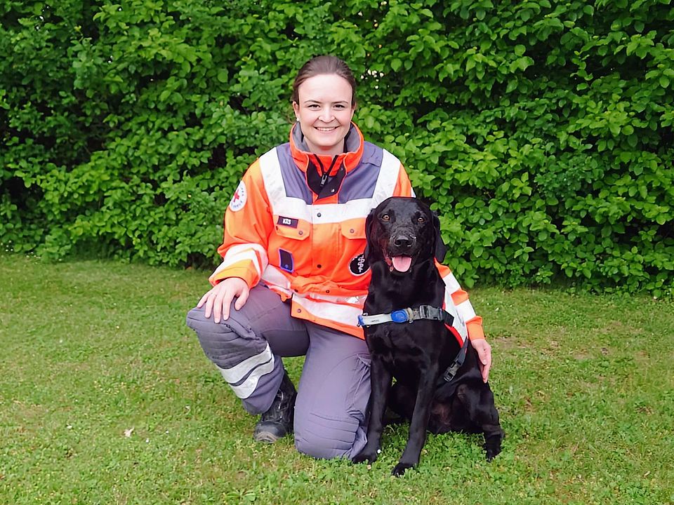 Lächelnde Rettungshundeführerin Eva in orange-grauer Einsatzkleidung kniet auf einer Wiese neben sitzendem, schwarzen Rettungshund Yari mit Geschirr; im Hintergrund dichter grüner Busch.