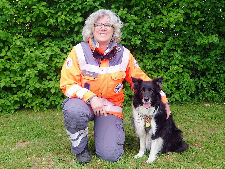 Lächelnde Rettungshundeführerin Anja in orange-grauer Einsatzkleidung kniet auf einer Wiese neben einem sitzenden, schwarz-weißen Rettungshund Cailin mit Geschirr; im Hintergrund dichter grüner Busch.