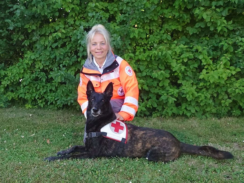 Lächelnde Rettungshundeführerin Christine in orange-grauer Einsatzkleidung kniet auf einer Wiese neben liegendem, schwarz-braunen Rettungshund Avventuro mit Geschirr; im Hintergrund dichter grüner Busch.