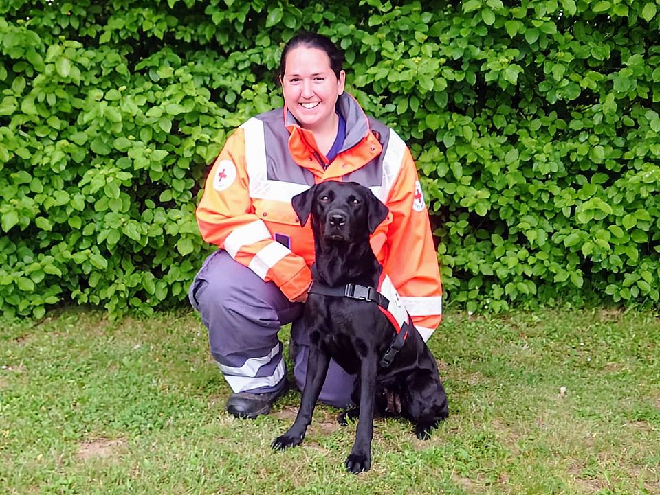 Lächelnde Rettungshundeführerin Saskia in orange-grauer Einsatzkleidung kniet auf einer Wiese neben einem sitzenden, schwarzen Rettungshund Bilyanu mit Geschirr; im Hintergrund dichter grüner Busch.